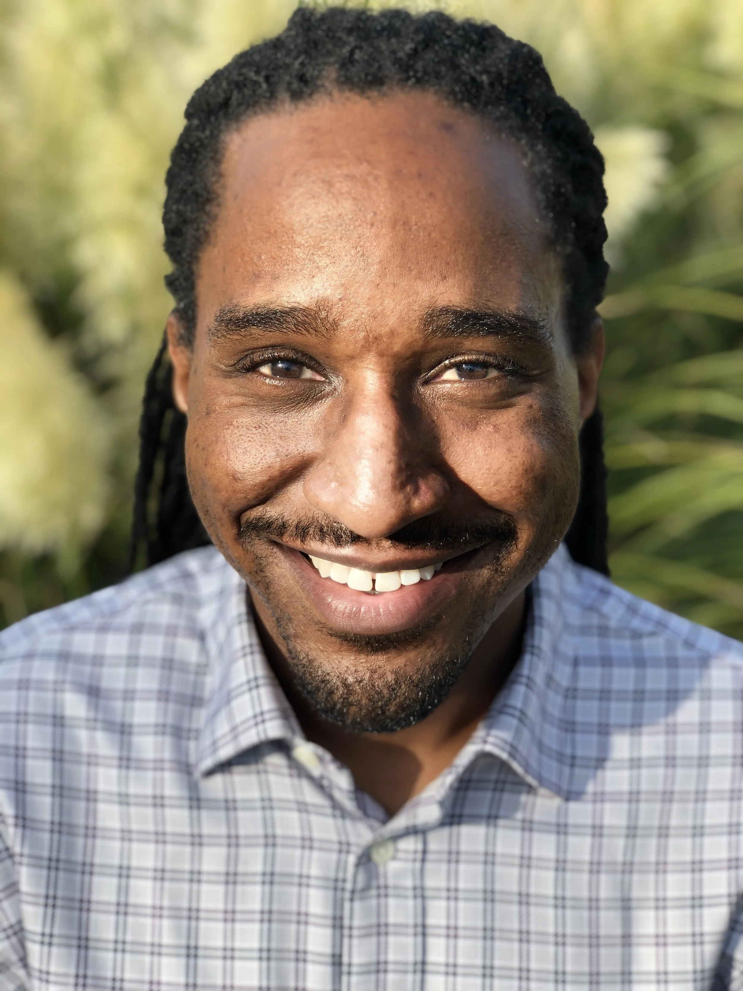 Close-up of a smiling man with dreadlocks, wearing a checkered shirt, outdoors with greenery in the background.