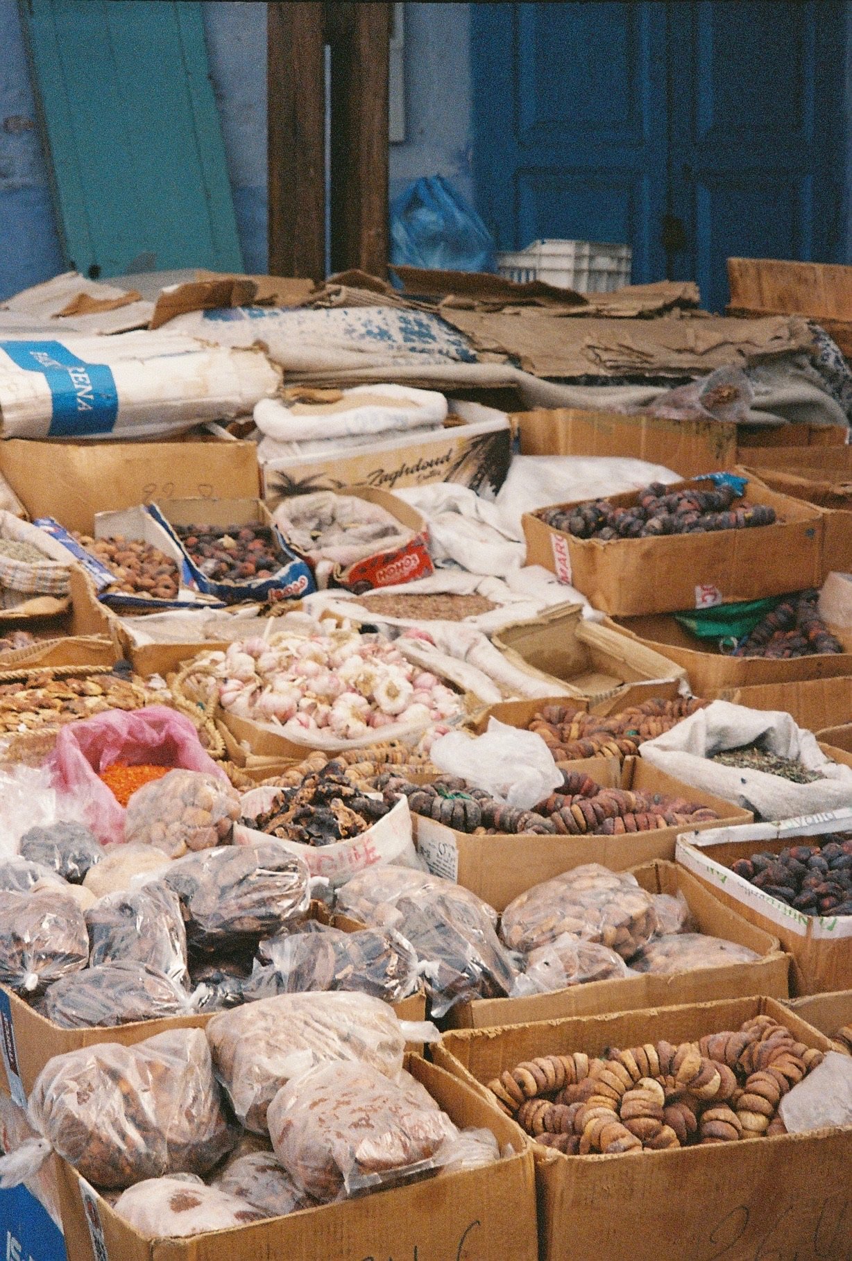 Various types of garlic, onions, and other dried produce displayed in open cardboard boxes at a market stall.