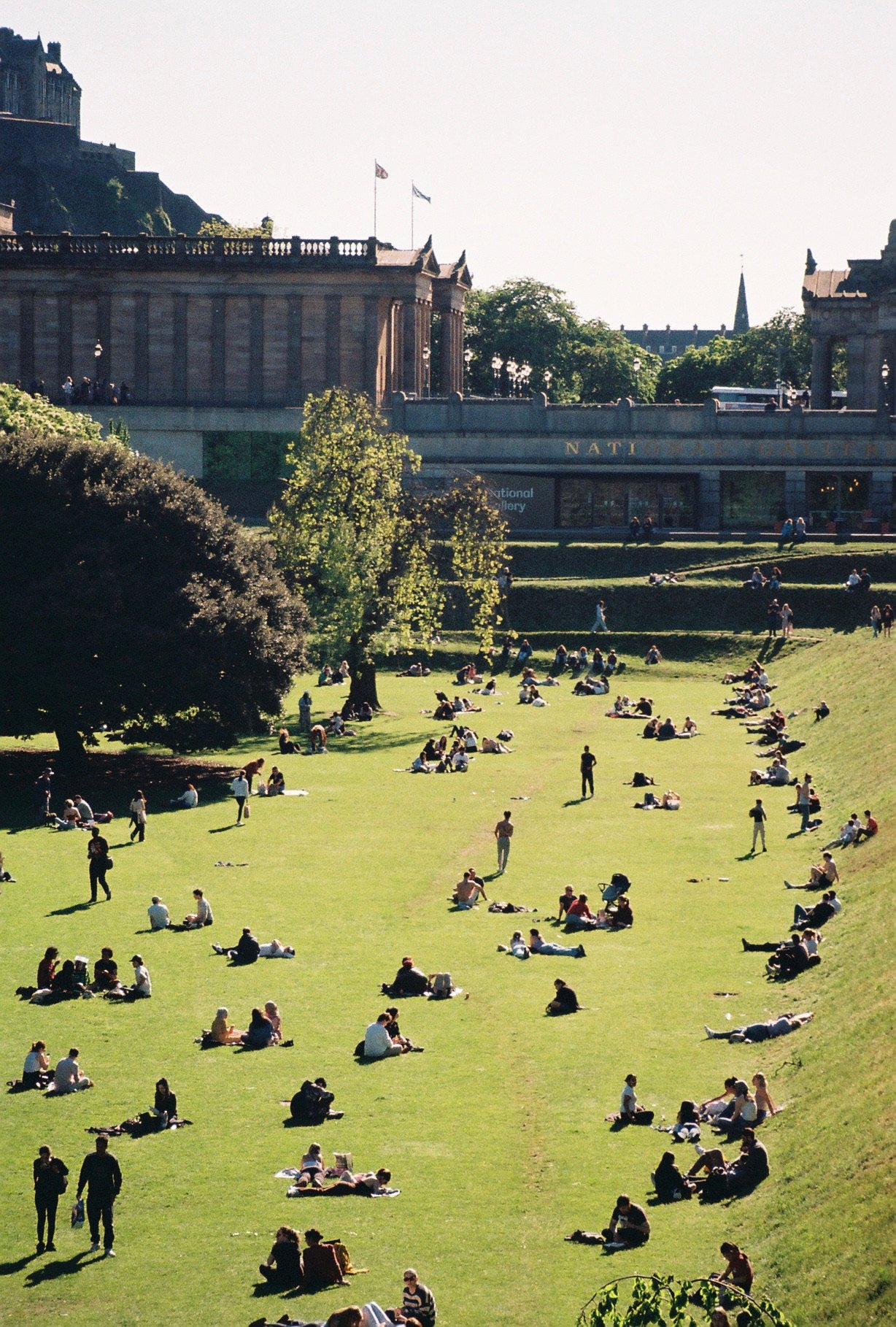 People relaxing and sitting on the grassy slope of a park in Edinburgh, Scotland, with buildings and trees in the background.