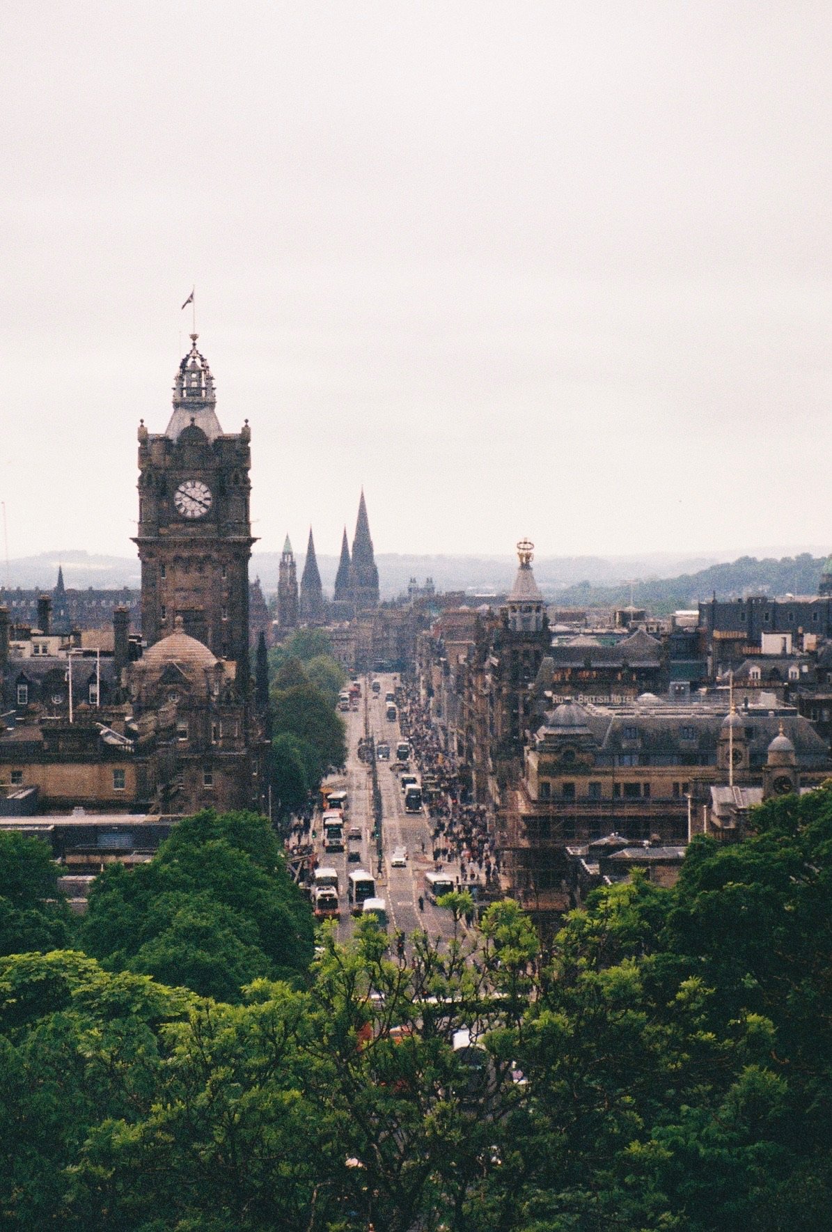 A city street scene in Edinburgh, Scotland, with the prominent clock tower and historic buildings, surrounded by green trees, under an overcast sky.