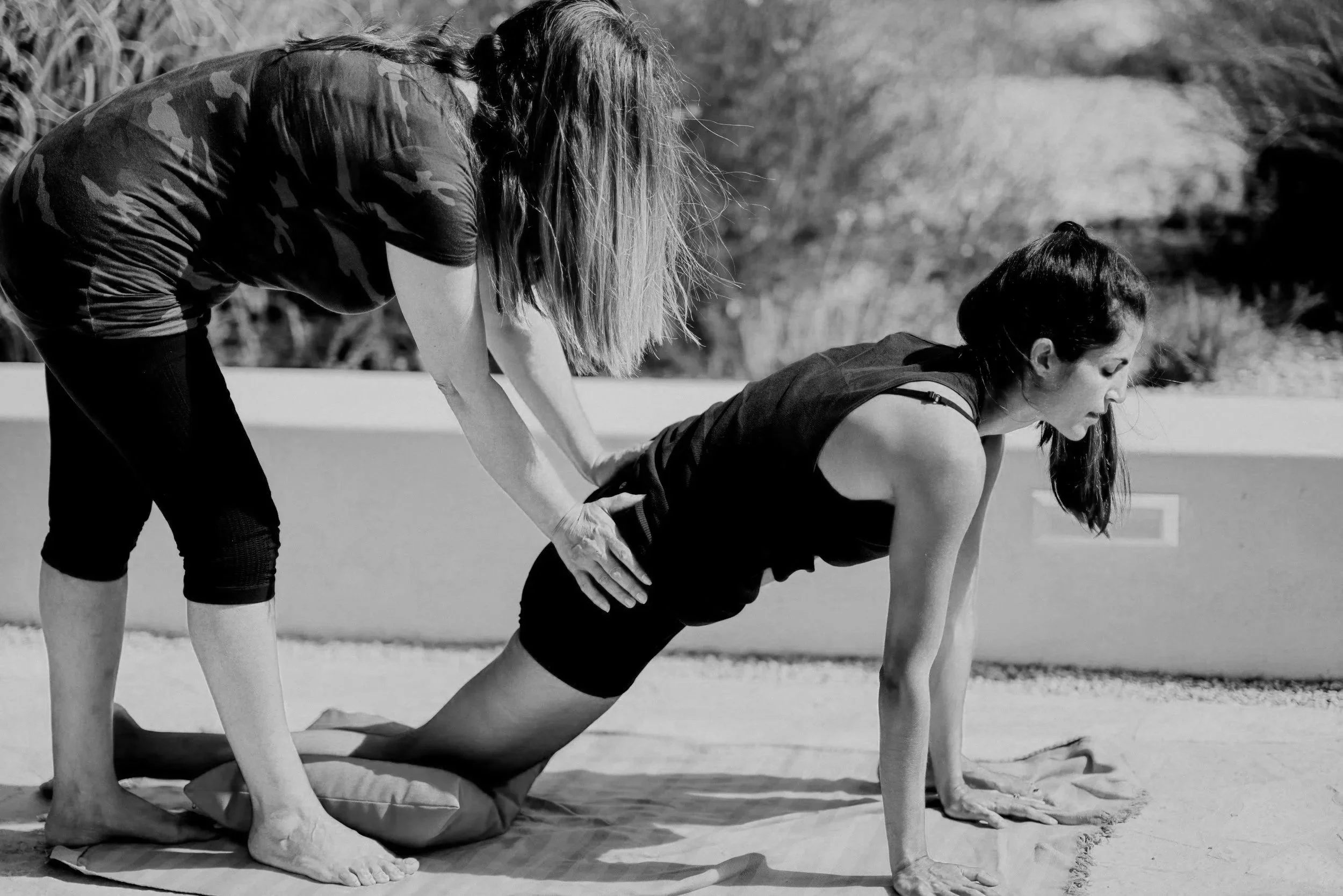 Two women practicing yoga outdoors, one in a kneeling position and the other in a forward bend, on a sunny day.