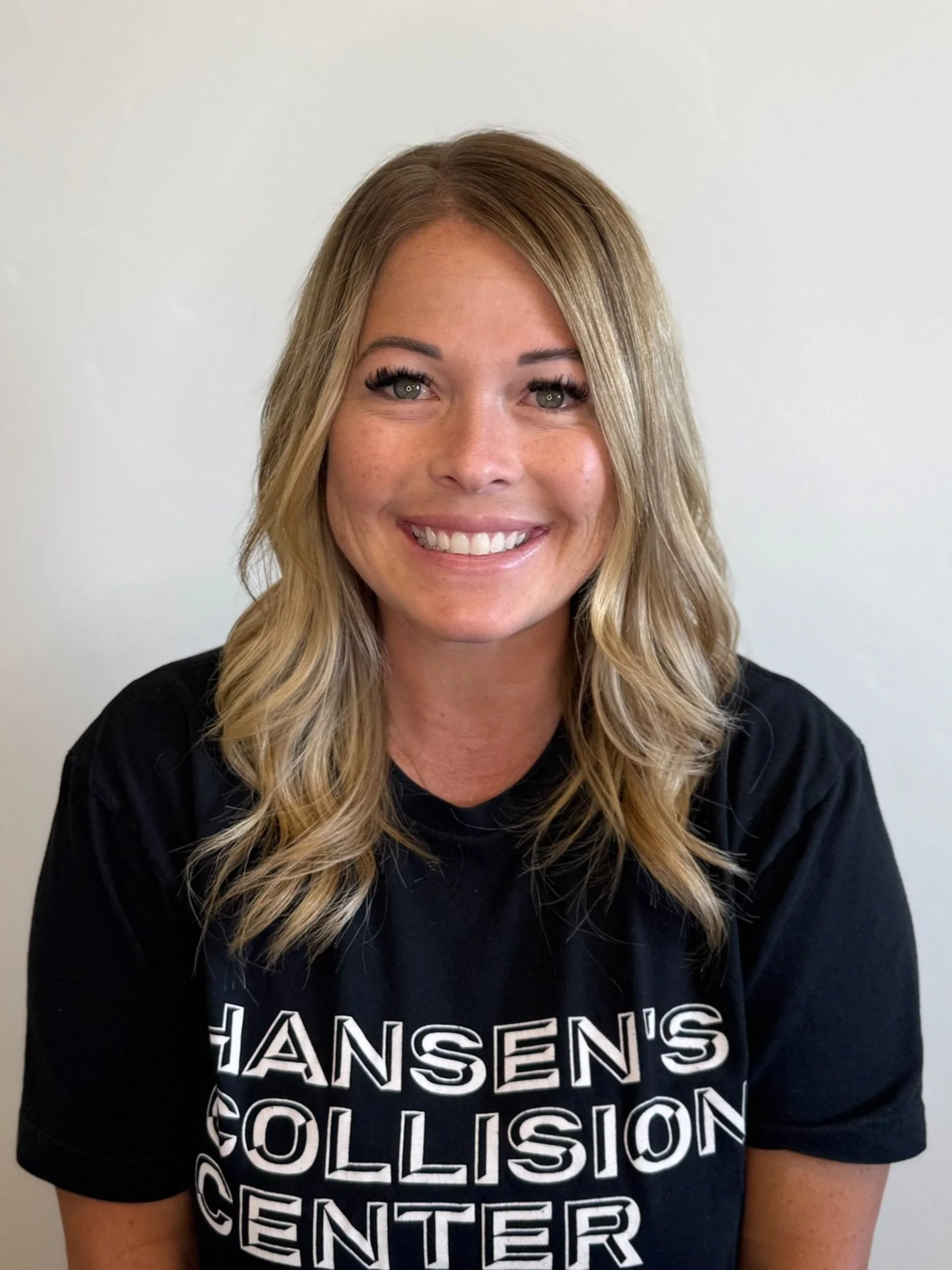 A woman with shoulder-length blonde hair, smiling, wearing a black t-shirt that reads 'HANSEN'S COLLISION CENTER'.