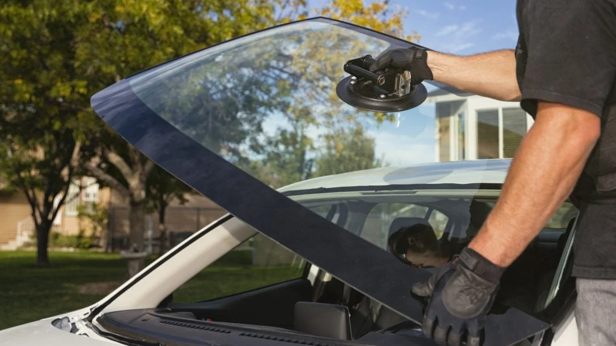 Person cleaning the windshield of a white car with a power sander, wearing black gloves, outdoors with trees and houses in the background.
