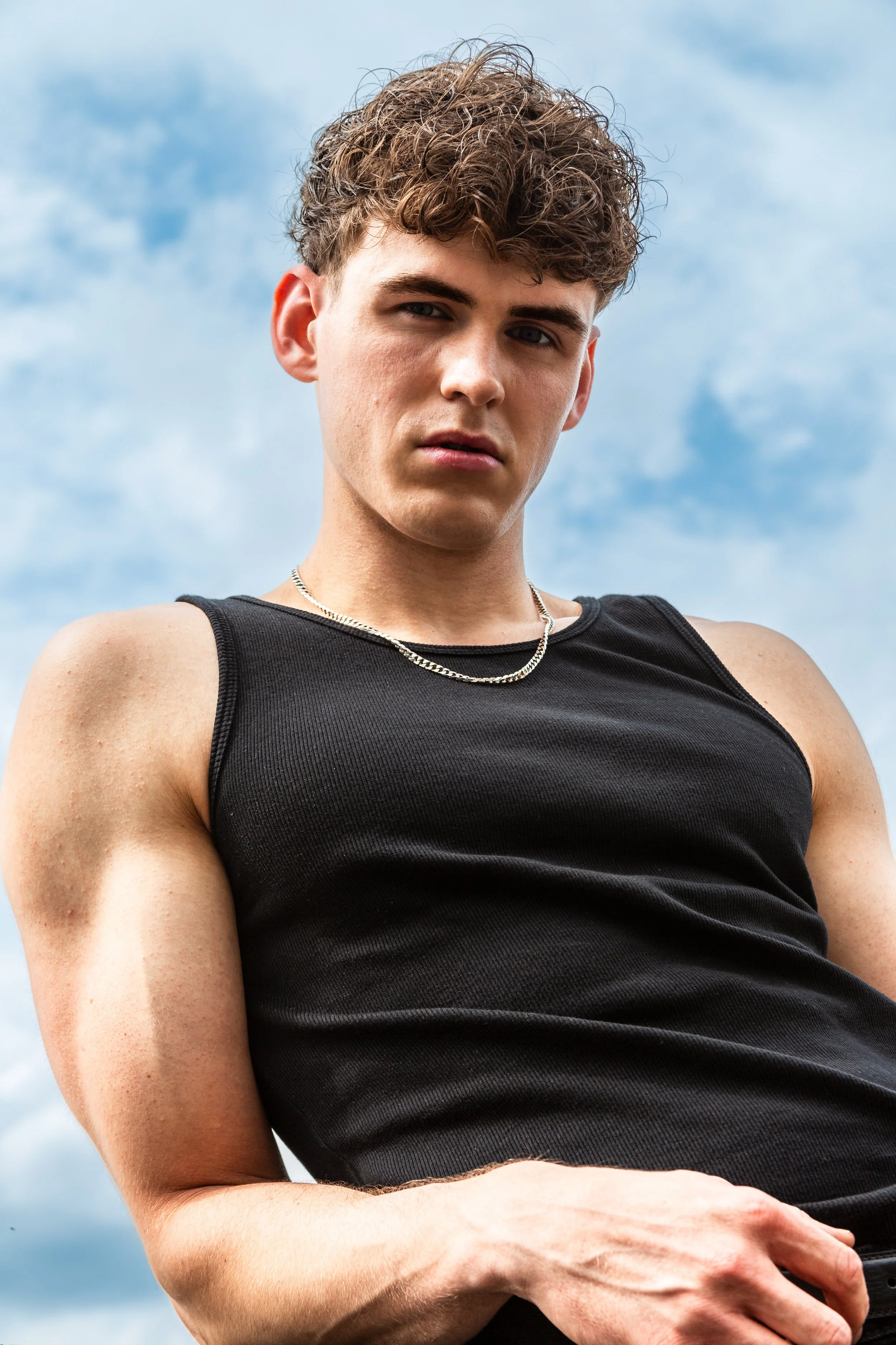 Young man with curly hair wearing a black sleeveless shirt and silver necklace outdoors against a blue sky with clouds.