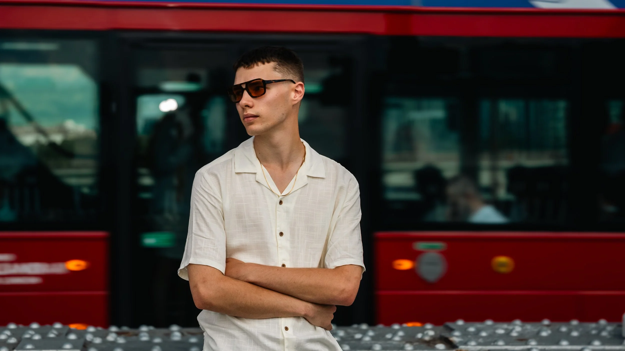 A young man with short dark hair and sunglasses standing with arms crossed in front of a red bus.