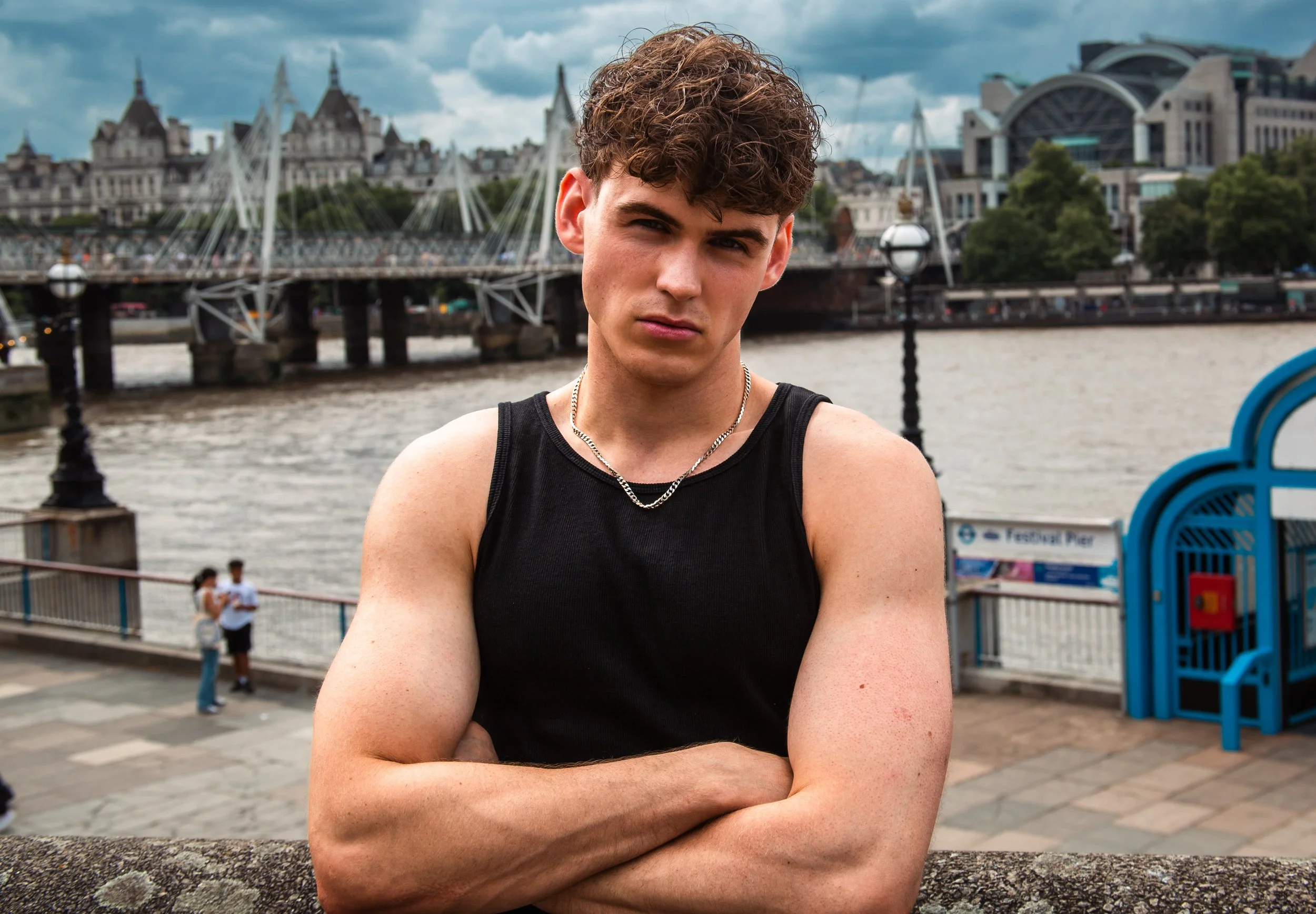 A young man with curly brown hair and fair skin standing outdoors near a river, crossing his arms, wearing a black sleeveless shirt and silver chain necklace, with a cityscape featuring a bridge and modern buildings in the background.