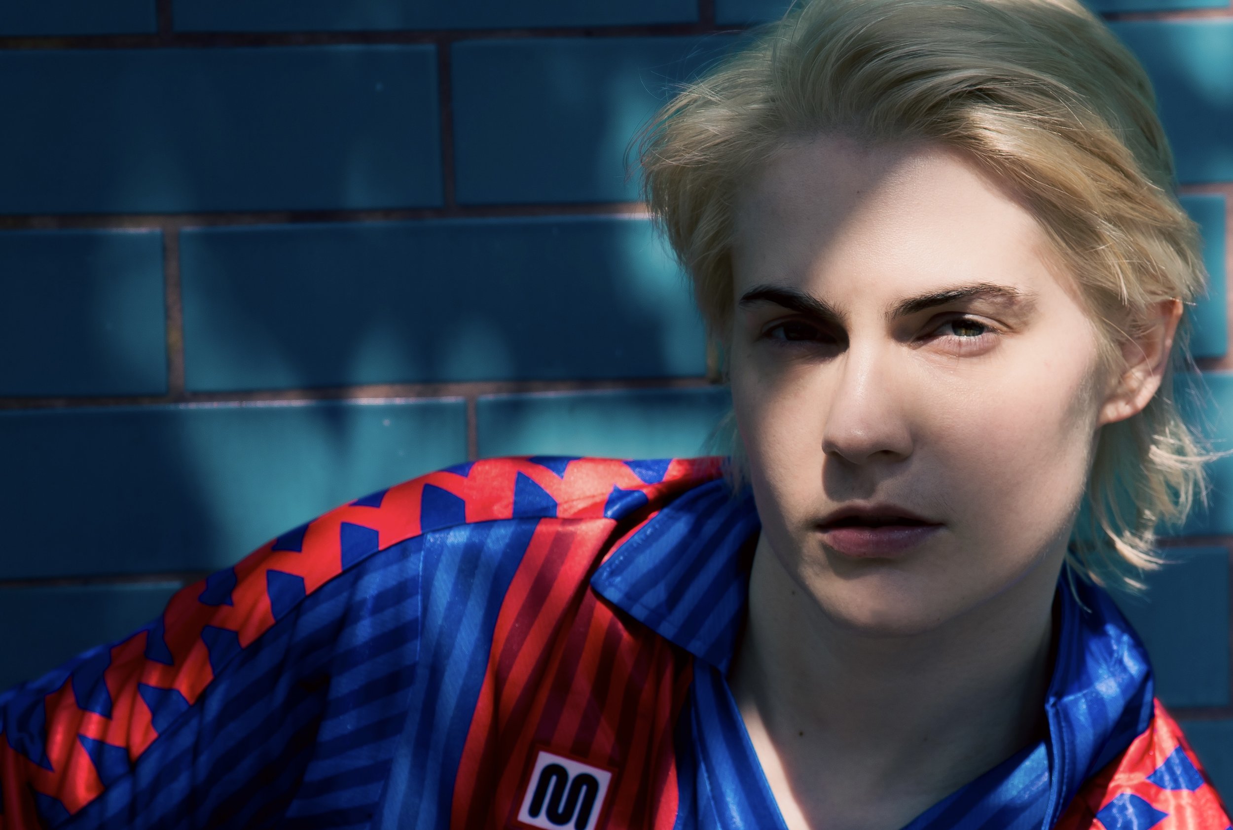A young woman with short blond hair and fair skin, wearing a blue and red sports jersey, leaning against a blue tiled wall.