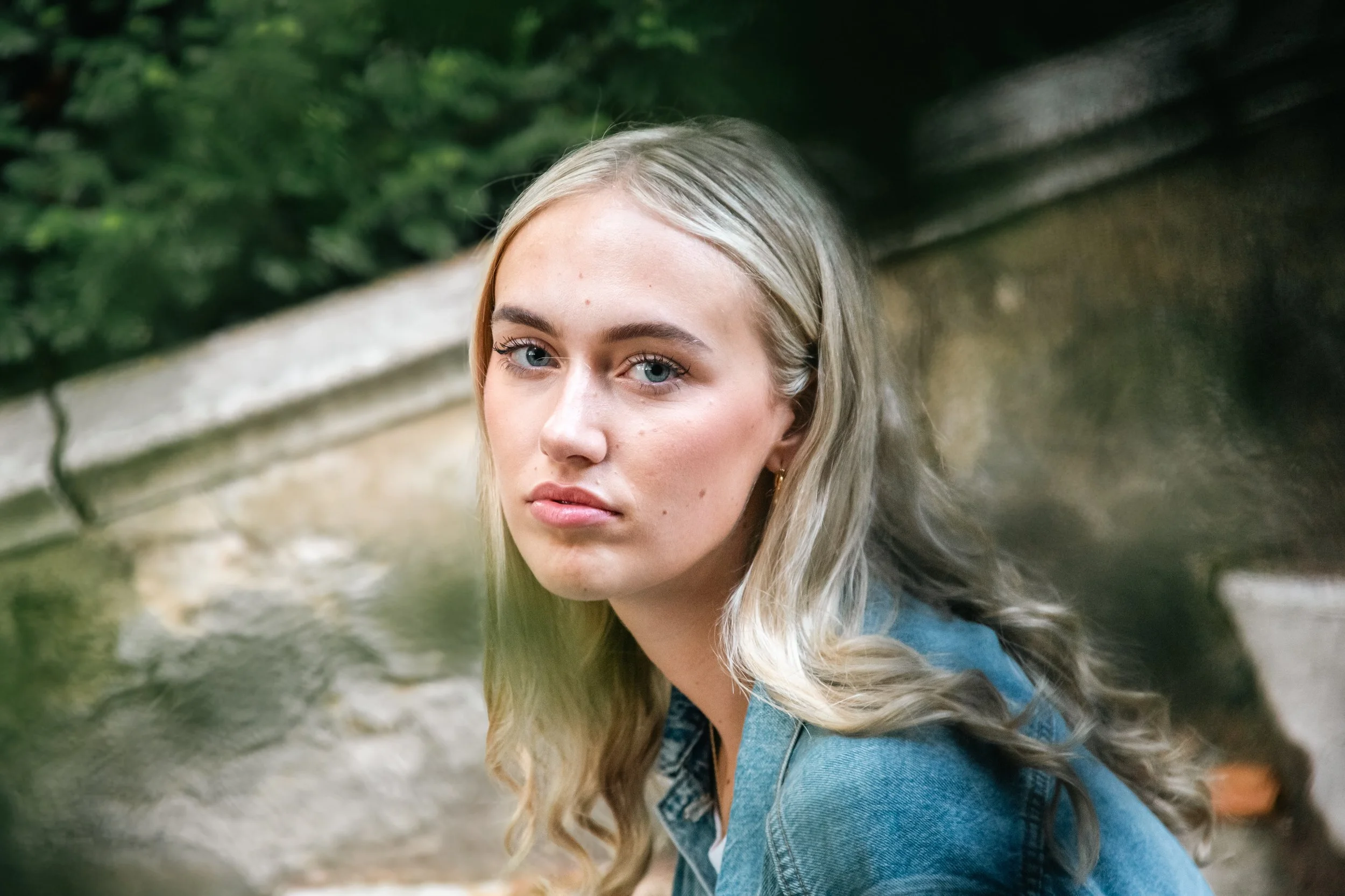 Portrait of a young woman with blonde hair and blue eyes, wearing a denim jacket outdoors near a stone water feature and green foliage.