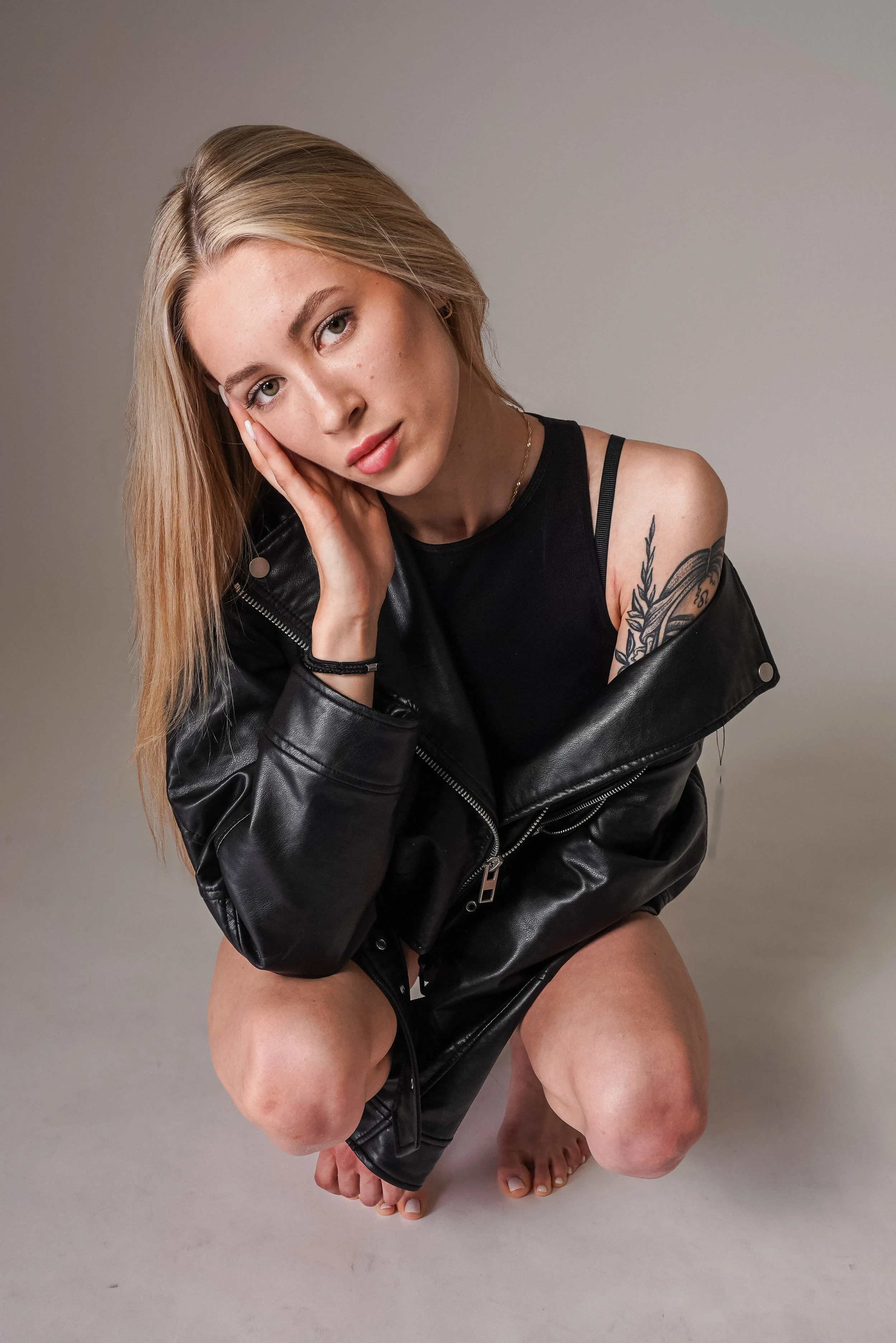A young woman with long blonde hair, wearing a black tank top and a black leather jacket, crouching barefoot with one hand resting on her face, looking directly at the camera against a neutral background.