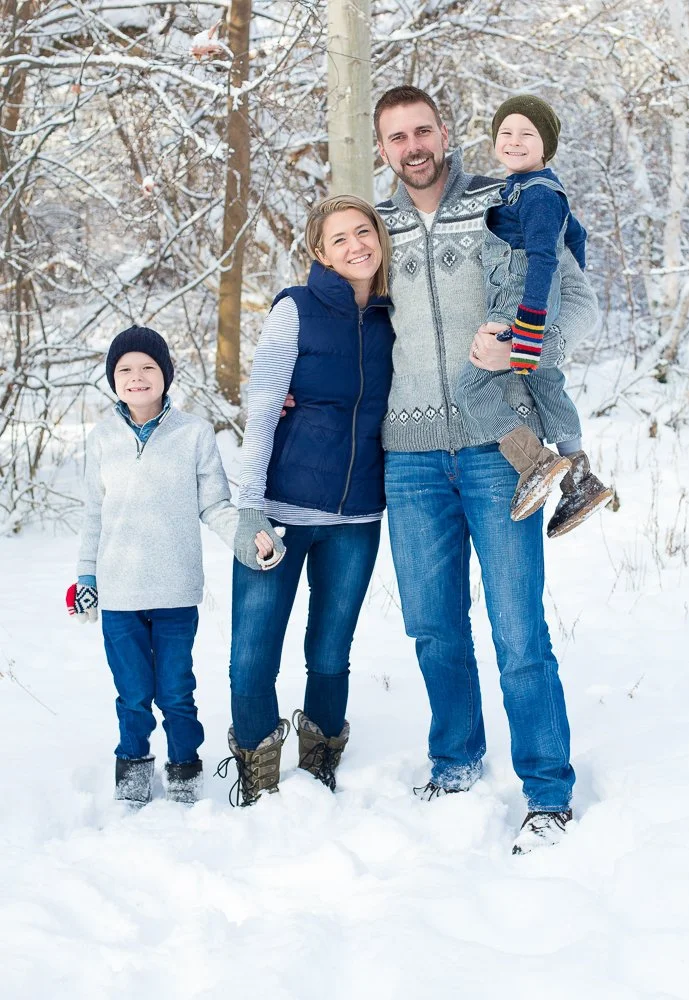 A family of four standing in snow-covered woods during winter, smiling and dressed in warm clothing.