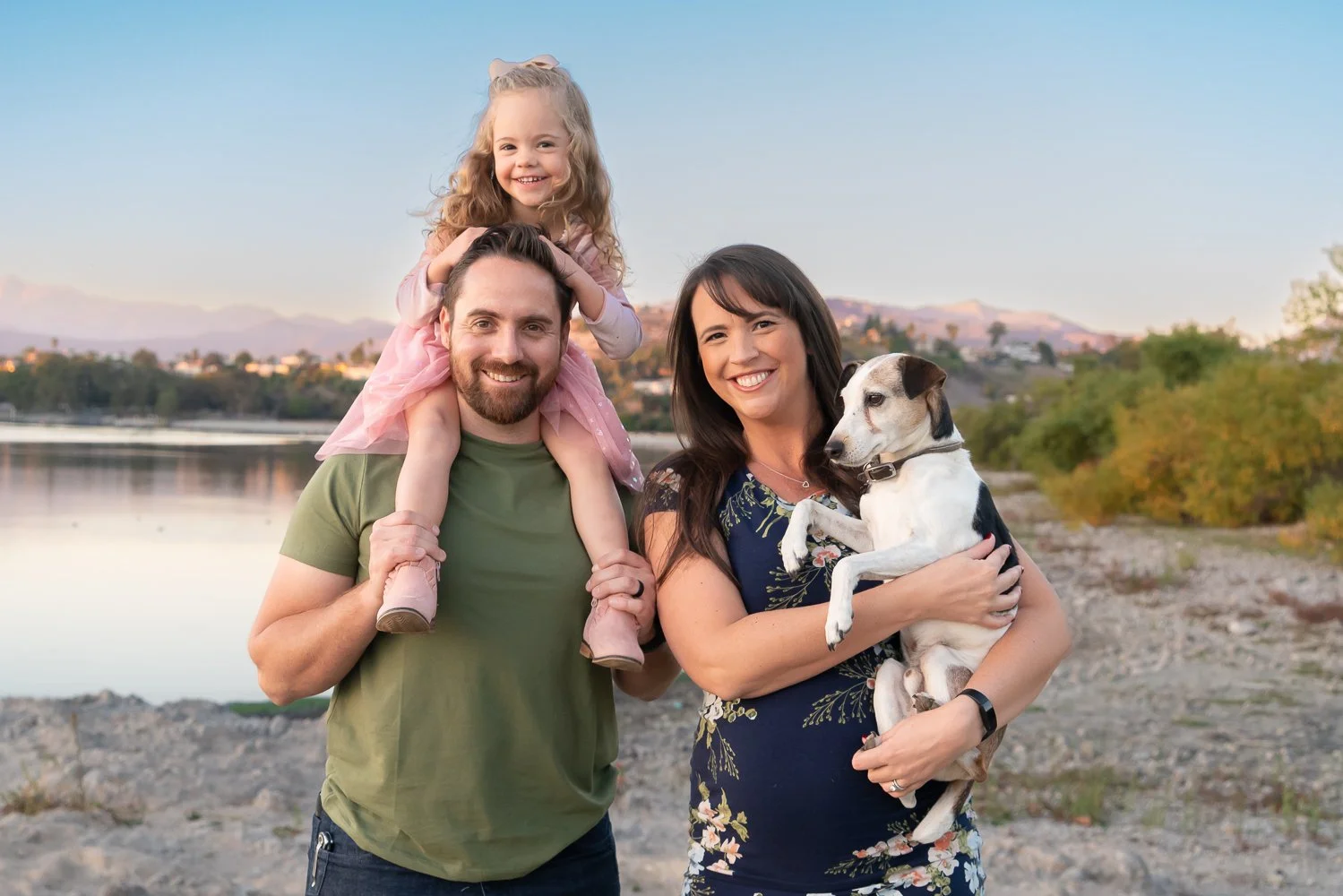 A family of three with a dog standing outdoors by a lake with hills in the background during sunset. The man is carrying a little girl on his shoulders, the woman holds the dog, and all are smiling.