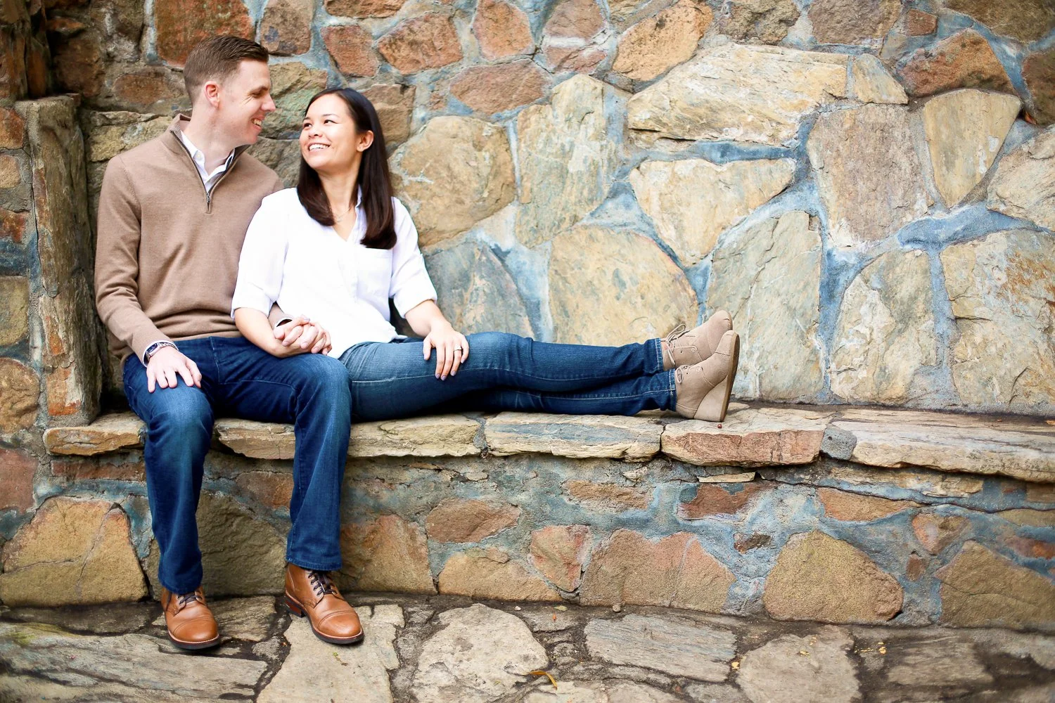 A couple sitting on a stone ledge against a stone wall, holding hands and smiling at each other.