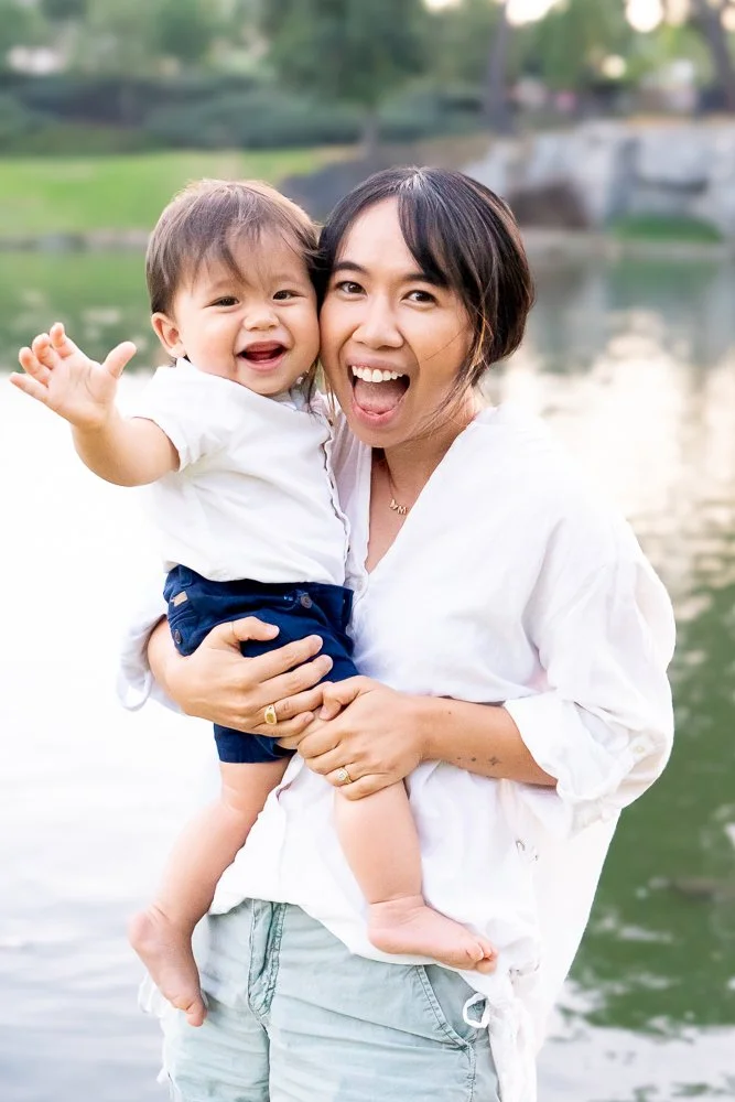 A woman holding a smiling toddler near a body of water outdoors.