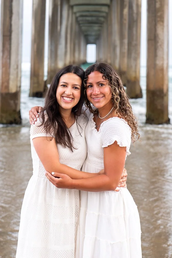 Two women in white dresses hugging under a pier at the beach, with ocean water around them.