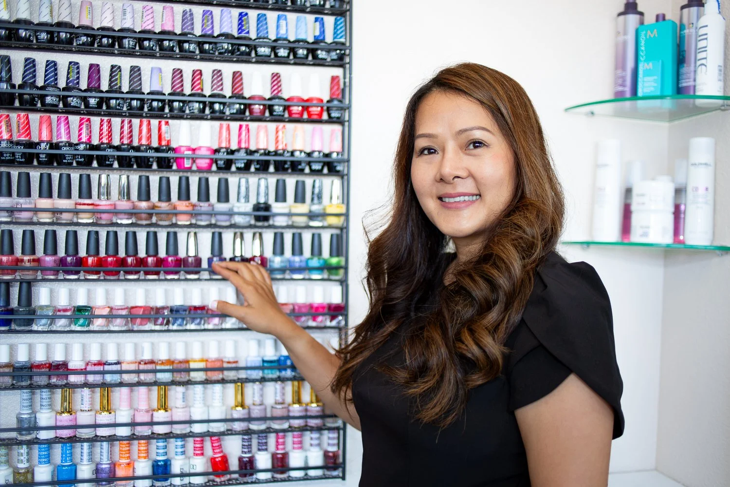 Woman with long brown hair selecting nail polish from a display rack filled with various colorful nail polish bottles.