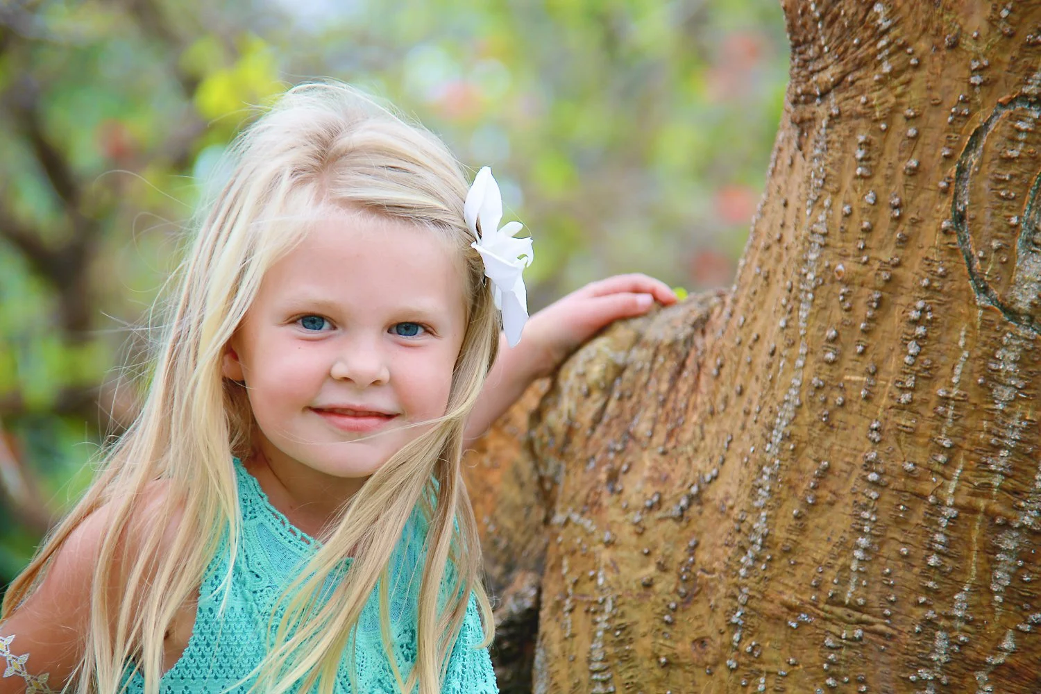 A young girl with long blonde hair, wearing a turquoise dress with lace, smiling and touching a tree in an outdoor setting with blurred green foliage in the background.