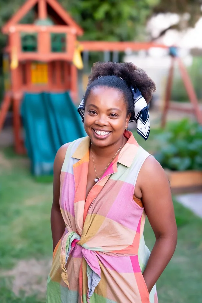 A smiling woman with dark curly hair tied back with a blue and white scarf, wearing a colorful sleeveless shirt, standing outdoors in front of a wooden playset with a slide.