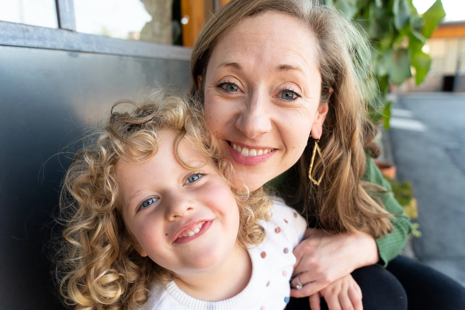 A woman and young girl smiling and leaning close together outdoors, sitting on a bench with green foliage in the background.
