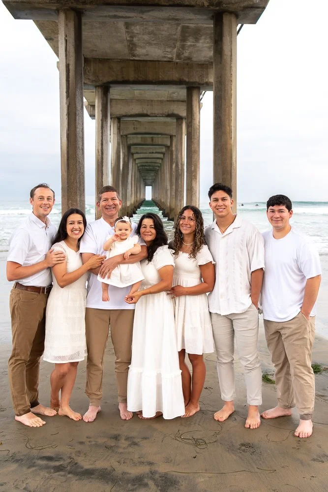 Group of eight family members in white and beige clothing standing on the beach under a pier with ocean in the background.