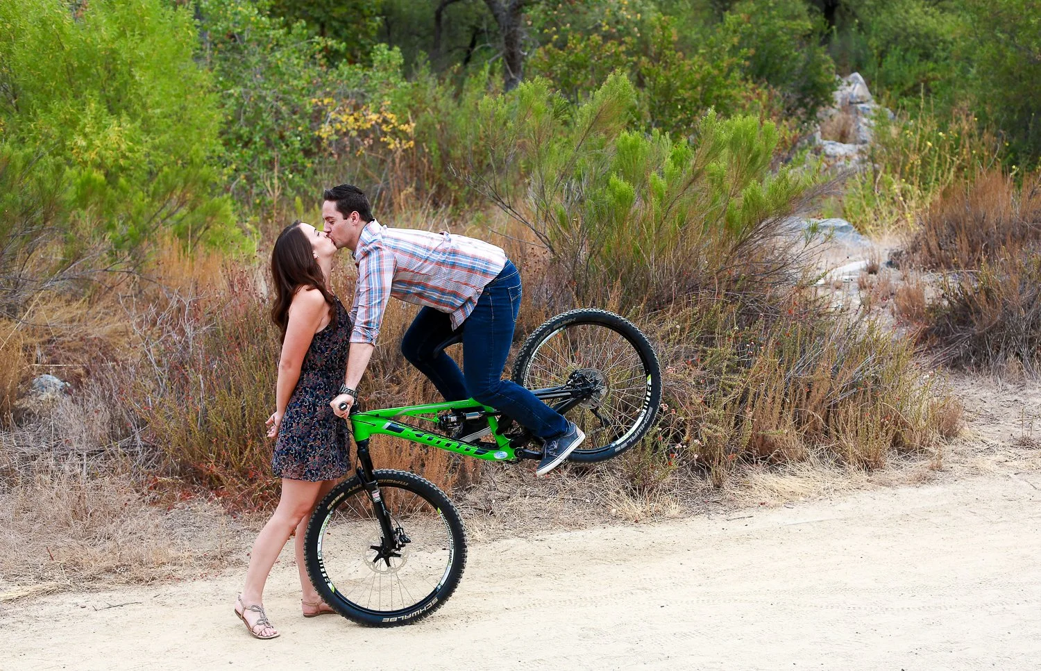 A man on a mountain bike and a woman are kissing on a dirt trail surrounded by natural vegetation and trees in a forested area.
