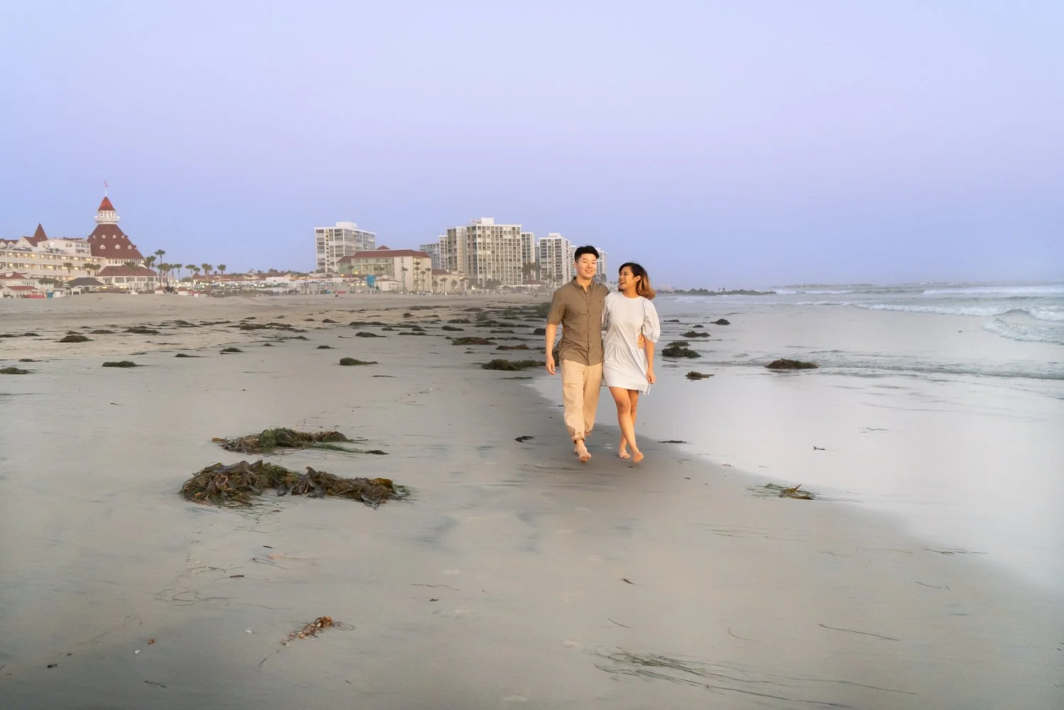 A couple walking barefoot on the beach with sand and seaweed, smiling and looking at each other, with city buildings and a castle-like structure in the background during sunset or early evening.