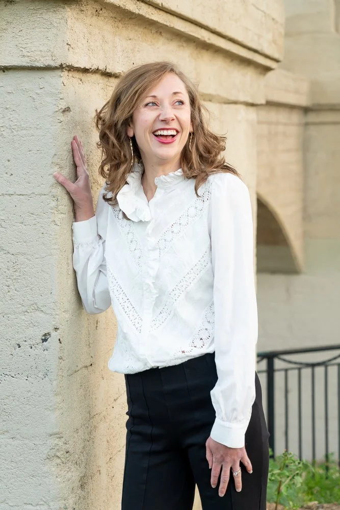 A woman with wavy, shoulder-length hair, laughing and leaning against a beige brick wall outdoors, dressed in a white blouse with ruffles and black pants.