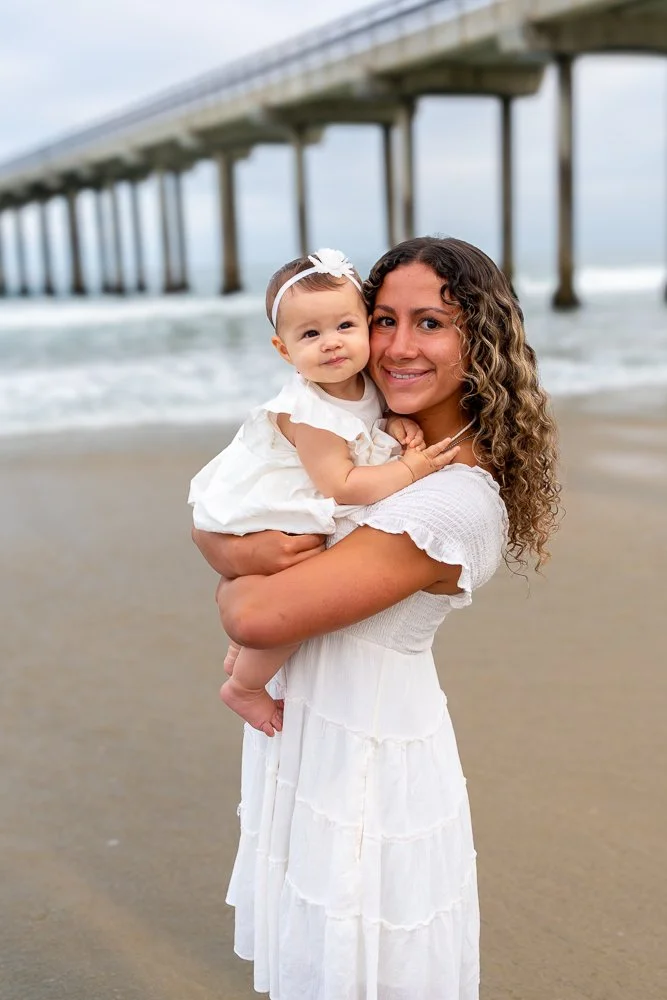 A woman holding a young girl on a beach with a pier in the background, both smiling and wearing white dresses.