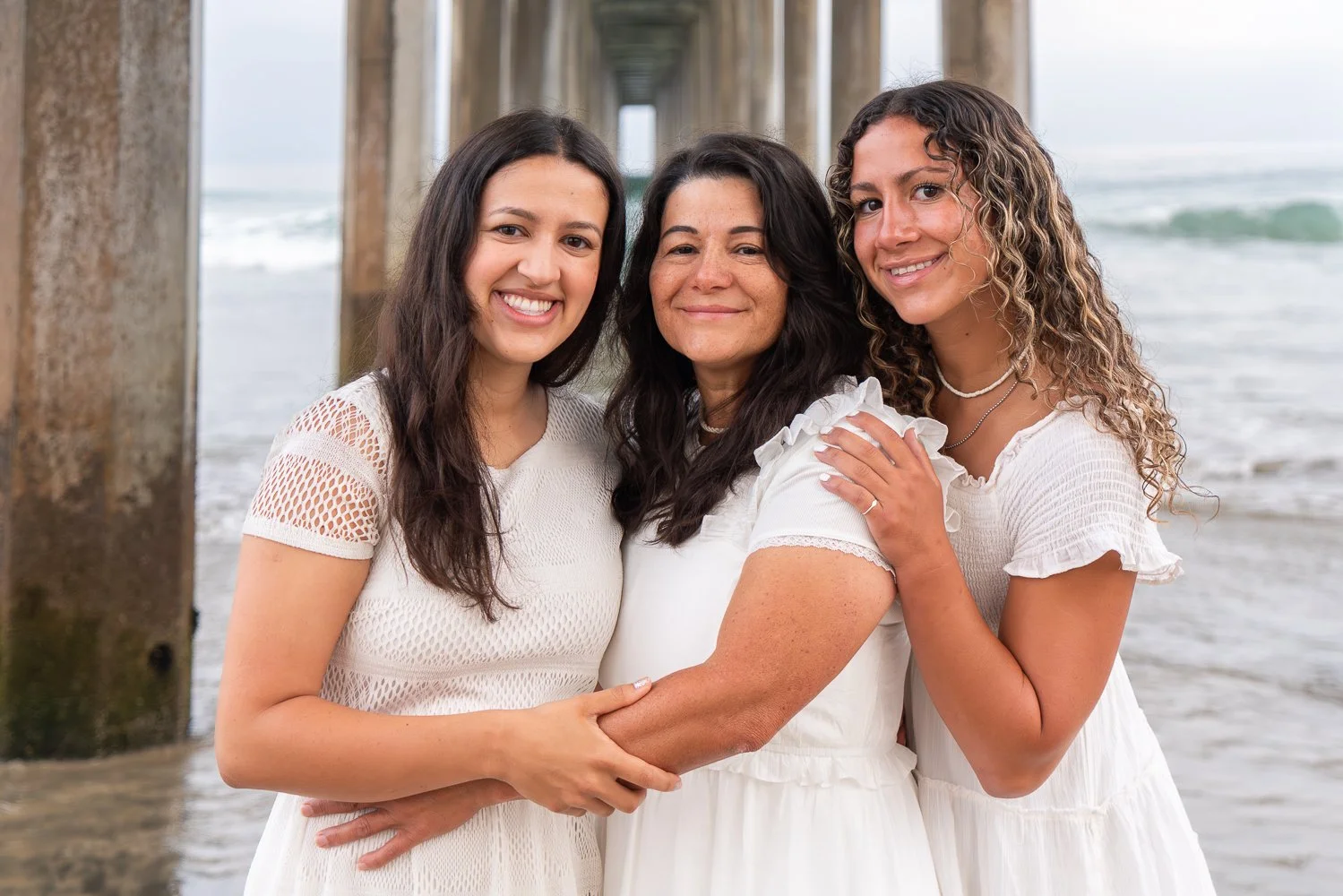 Three women smiling and embracing under a pier at the beach, wearing white dresses.