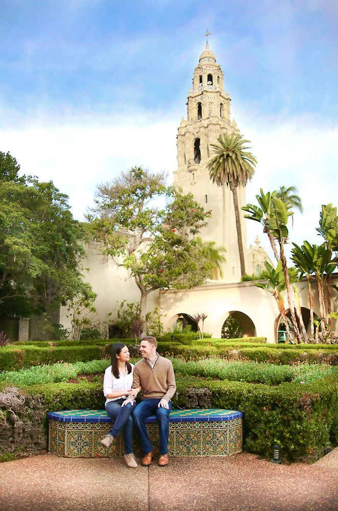 A couple sitting on a decorative tiled bench in a lush garden with a historic tower in the background, likely the Old Courthouse in San Diego.