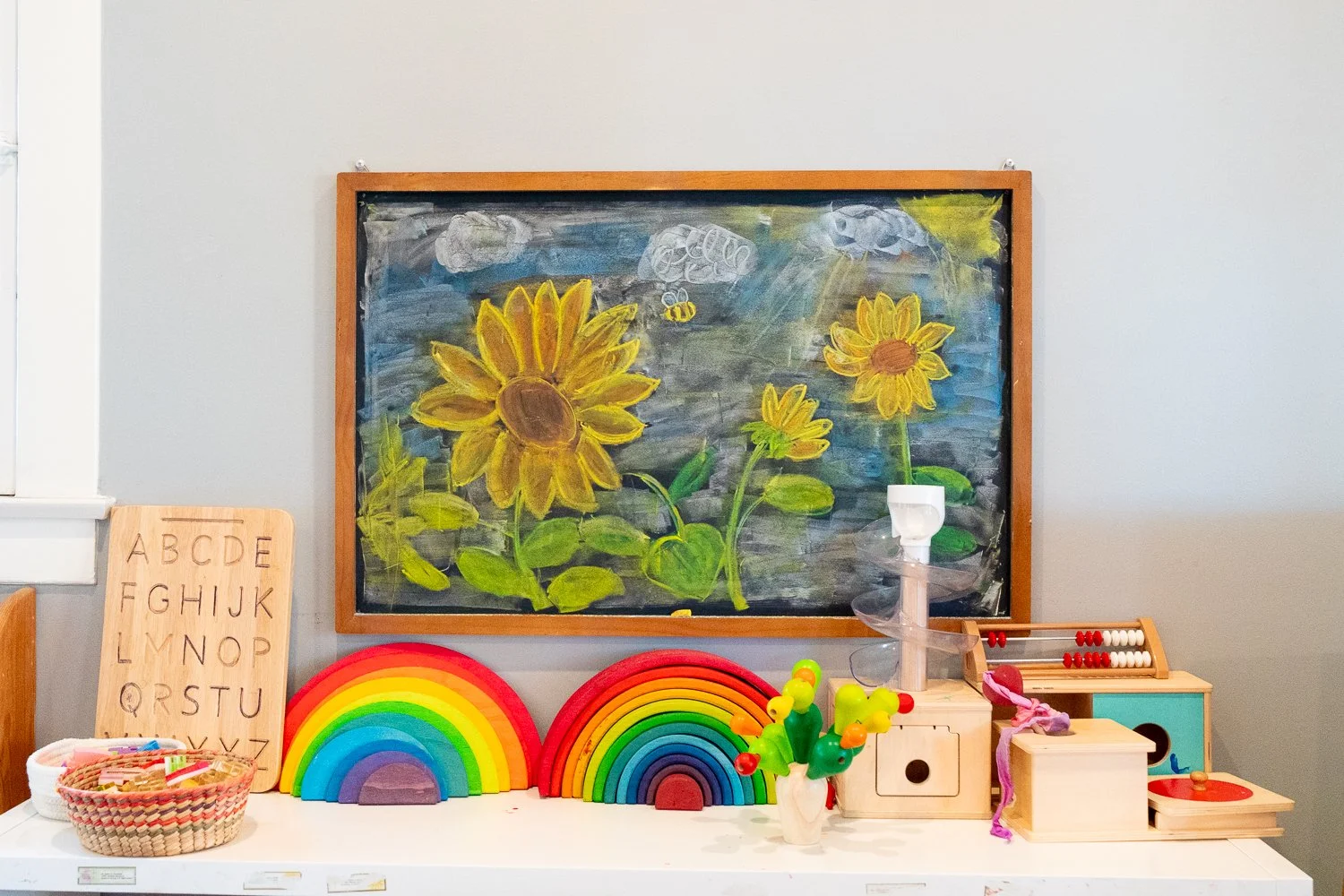 A classroom shelf with educational toys and decorations, a colorful rainbow decoration, a potted plant with yellow and red flowers, and a blackboard with a painting of sunflowers and a bee.