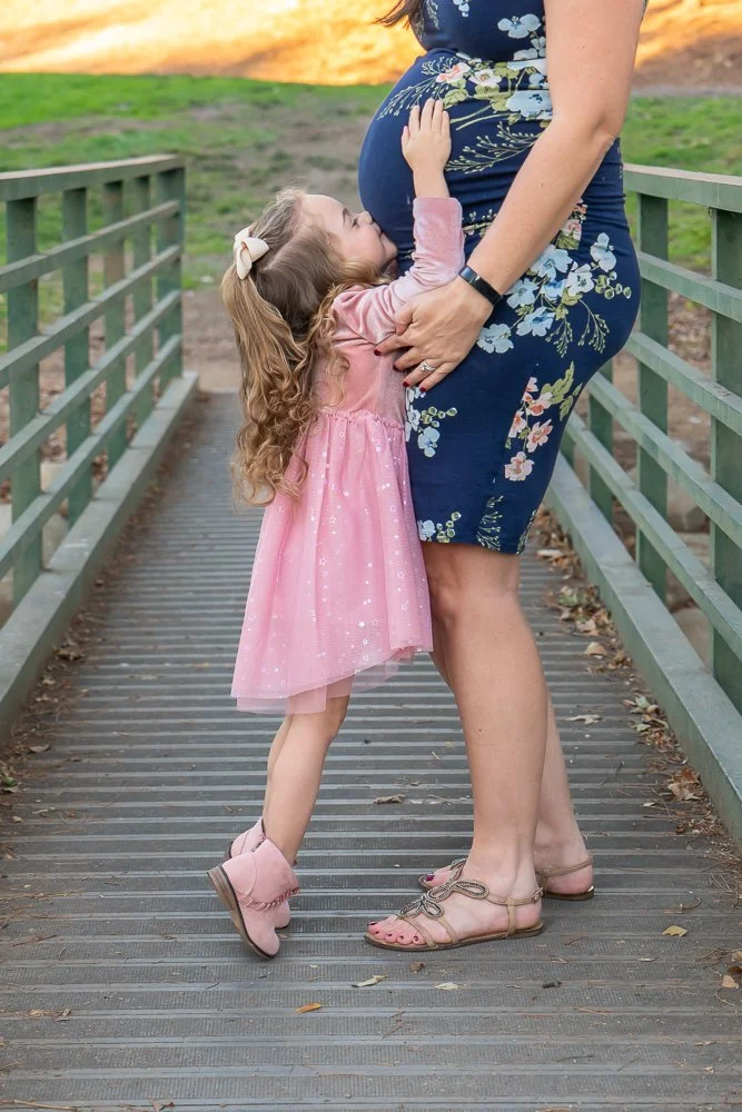 A young girl in a pink dress hugging a pregnant woman on a bridge, with the girl kissing the woman's belly.