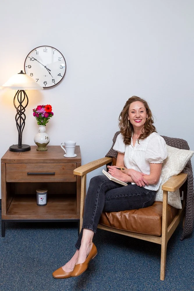 A woman with curly hair and a white blouse sitting in a wooden armchair, holding a notebook and pen, smiling at the camera. Next to her is a small wooden side table with a white lamp, a vase of pink and red flowers, a white mug, and a candle. Behind