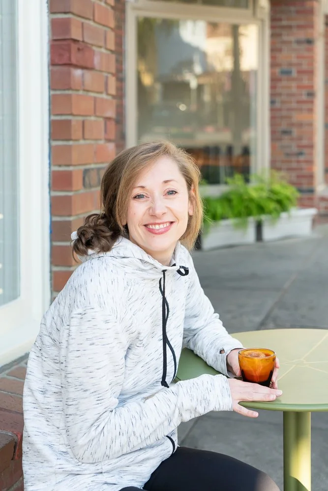 A woman with light brown hair and a white hoodie with black drawstrings sitting outdoors at a green table, holding a small amber glass, smiling at the camera, with a brick building and a window with a flower box in the background.