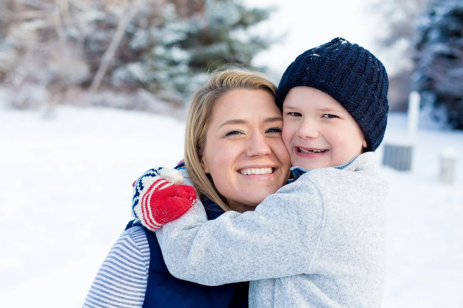 A woman and a young boy hugging outdoors in a snowy landscape, both smiling.