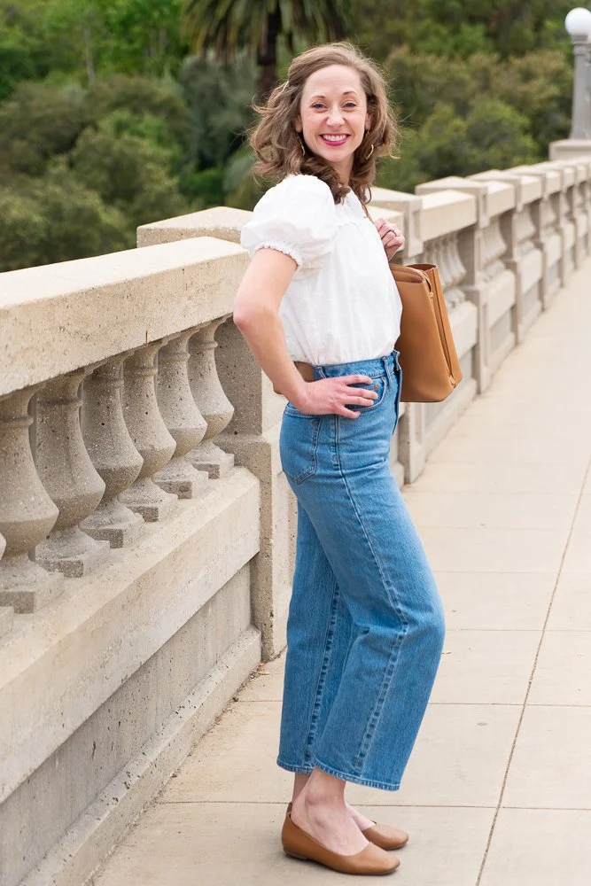 A woman with curly brown hair wearing a white puff-sleeve blouse, high-waisted blue jeans, and tan flats, standing next to a stone balustrade on a walkway surrounded by greenery and trees.