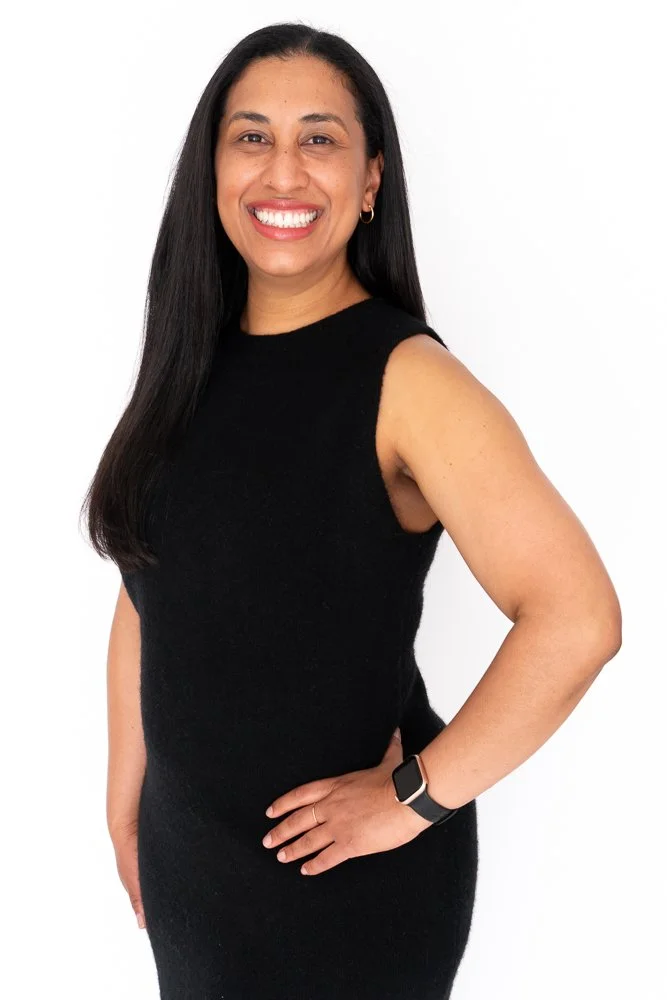 A woman with long black hair, wearing a sleeveless black dress and a smartwatch, smiling and standing with one hand on her hip against a white background.