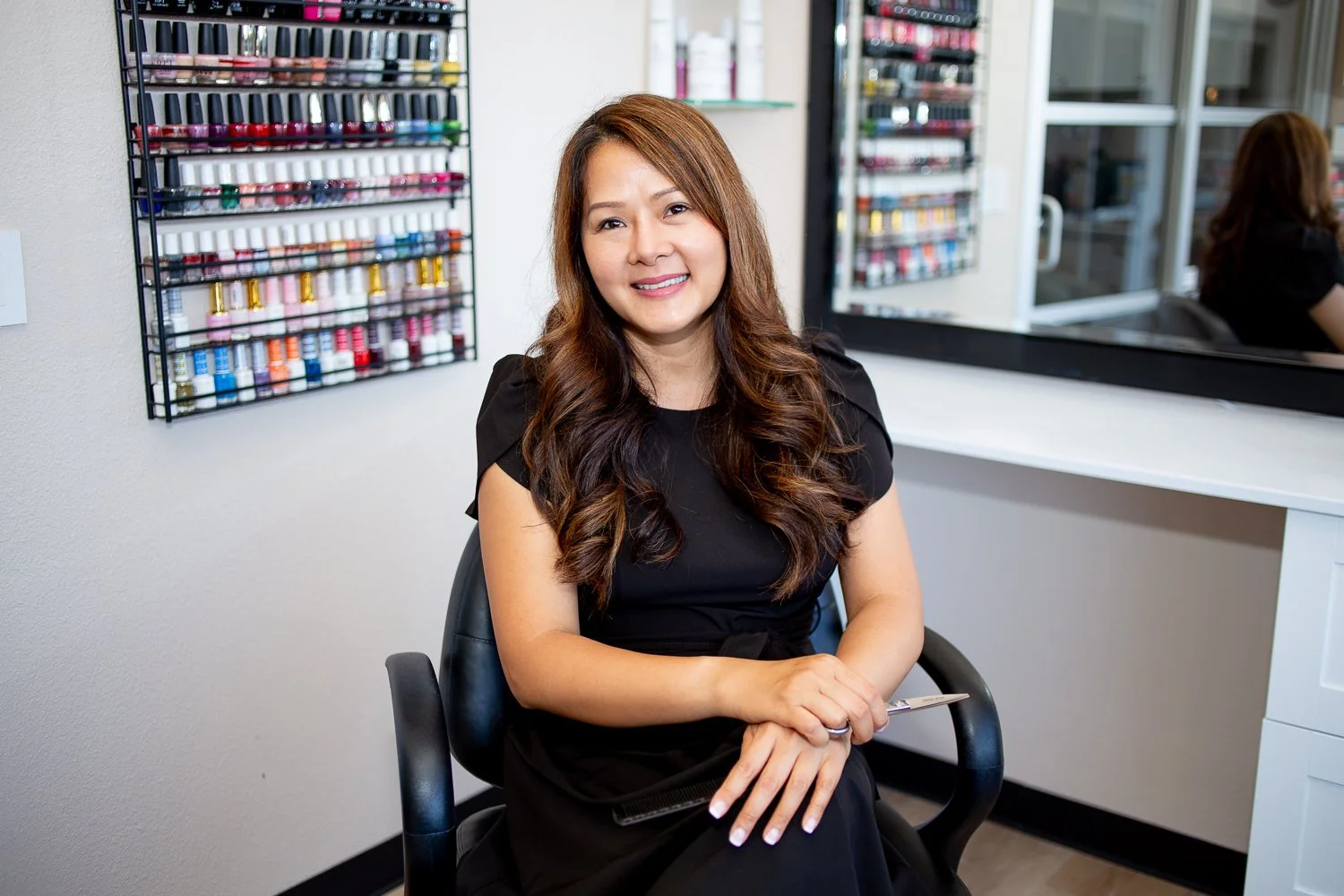 A woman with long, wavy brown hair smiling and sitting in a nail salon, with shelves of nail polish bottles visible behind her and a mirror reflecting another person in the background.