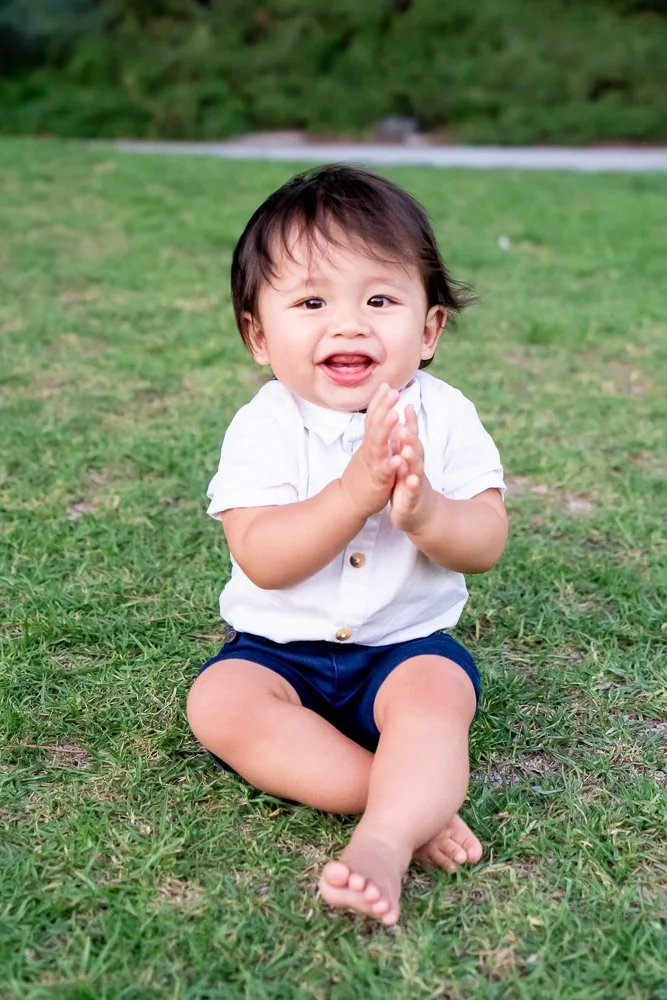 A young child sitting on grass outdoors, clapping hands, smiling, with trees in the background.