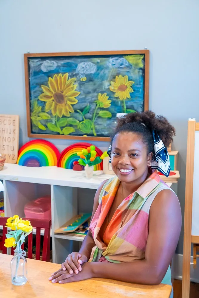 A woman smiling and sitting at a wooden table in a colorful classroom. There is a vase with yellow flowers on the table, and behind her are rainbow decorations, a chalkboard with a sunflower and cloud painting, and educational materials.