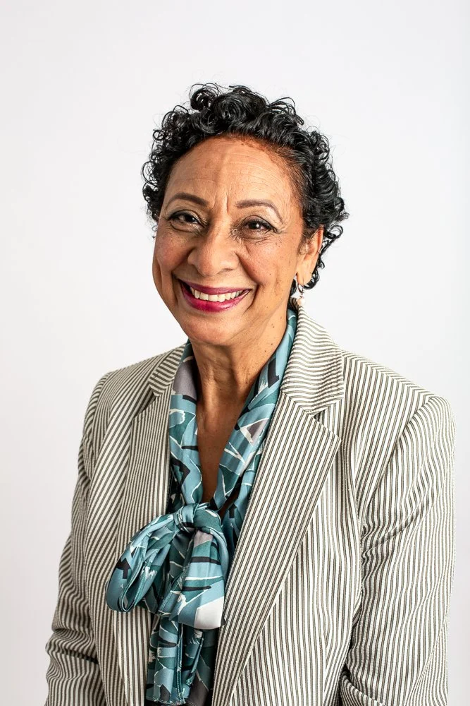 A woman with curly brown hair, smiling, wearing a yellow short-sleeve blouse with ruffled sleeves and blue jeans, standing outdoors on a concrete balcony with a scenic green landscape in the background.