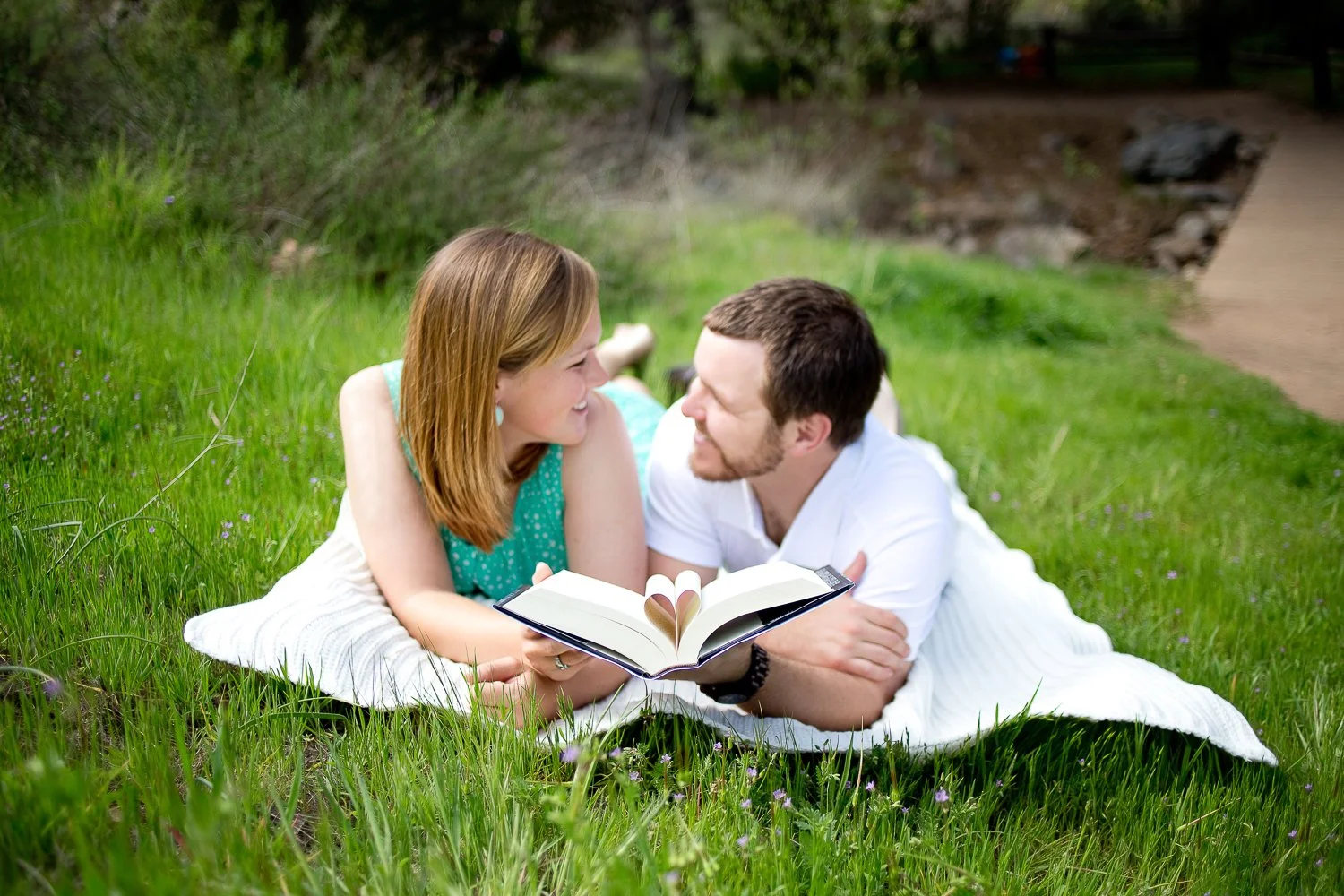 A young couple lying on a grassy field outdoors, sharing a book with a heart-shaped cutout in the pages, smiling and looking at each other.