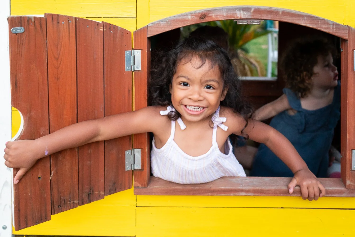 A young girl with curly hair smiling and leaning out of a decorated yellow playhouse window, with a boy behind her inside the playhouse.
