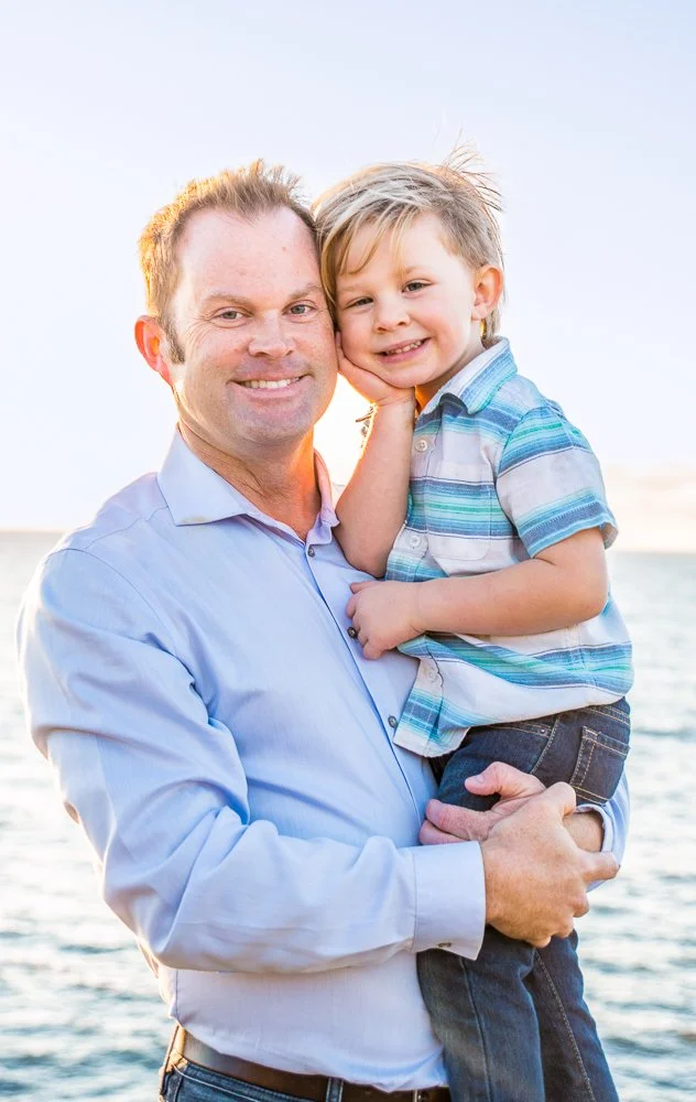 A man holding a young boy, both smiling, near a body of water during sunset.