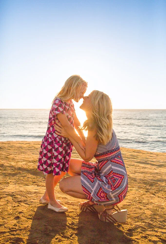 A woman and a young girl share a tender moment on the beach at sunset, with the woman kneeling and the girl standing close, touching noses.