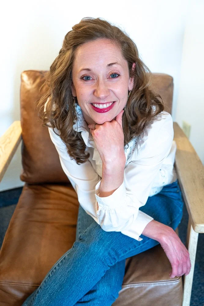 A woman with light skin and wavy, shoulder-length brown hair sitting on a brown leather couch with wooden armrests, smiling and resting her chin on her hand.