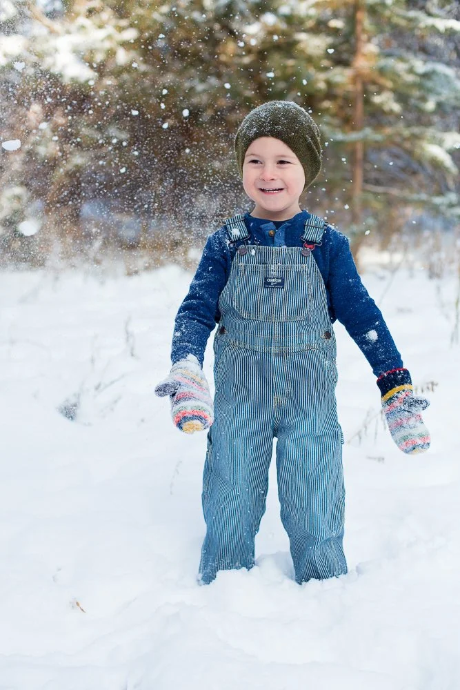A young boy playing in the snow, wearing a dark knit beanie, striped snow overalls, a blue long-sleeve shirt, and colorful mittens, with snow falling around him.