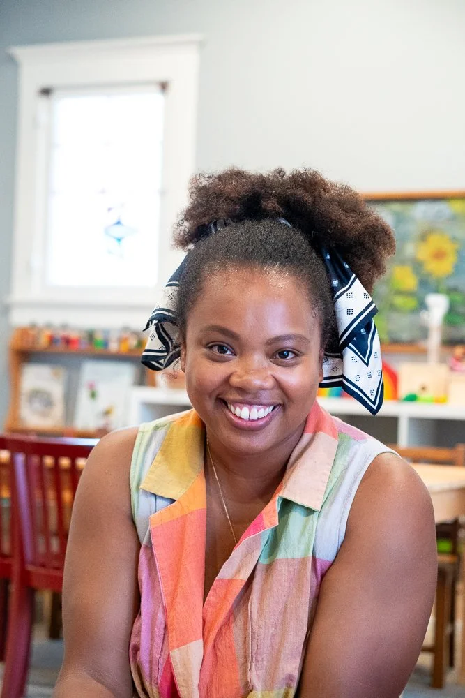 A smiling woman with curly hair tied back and a colorful scarf in a classroom or creative space.