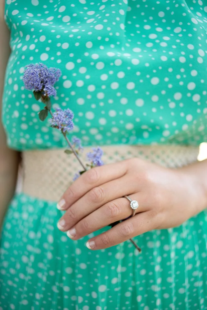 A woman wearing a teal dress with white polka dots is holding a purple flower and wearing an engagement ring with a large center diamond.