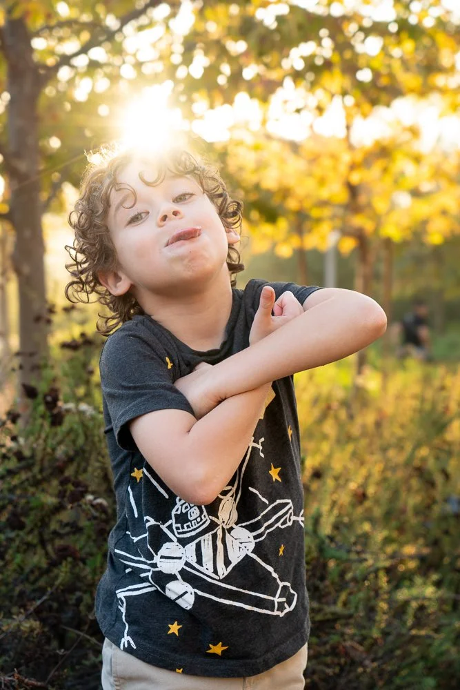 Young boy with curly hair standing outdoors in a wooded area during fall, with sunlight shining behind him. He is wearing a black T-shirt with a space-themed graphic and stars, posing with arms crossed.