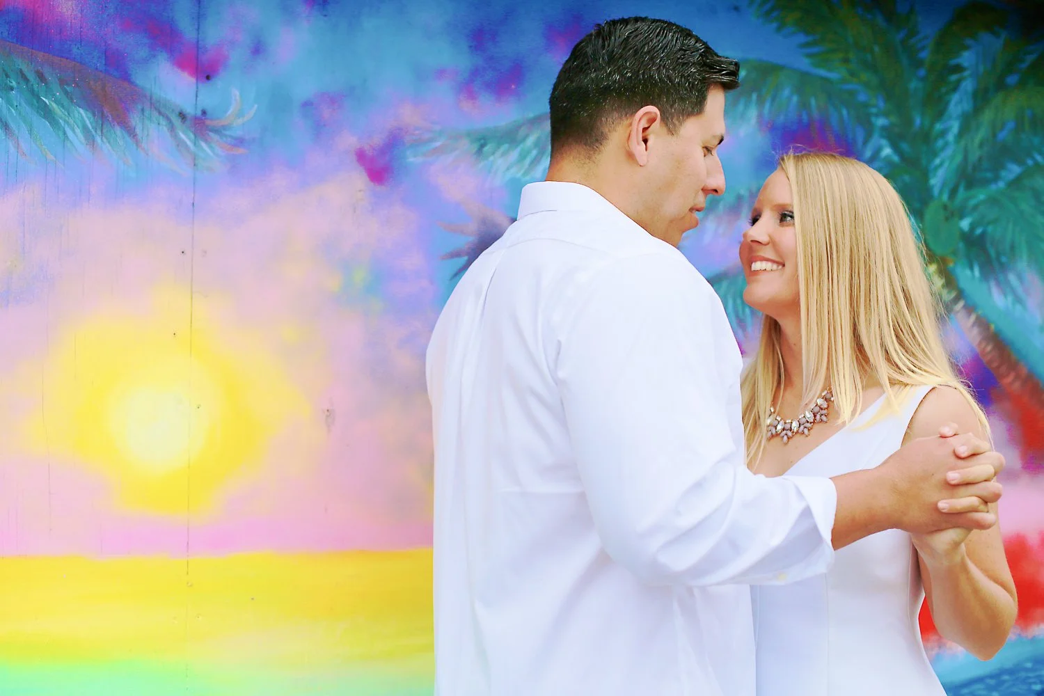 A man and woman holding hands and smiling at each other in front of a colorful abstract background with a sunset and palm trees.